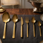 Luxury 36-piece gold-finished cutlery set featuring a geometric Greek key handle design, displayed elegantly on a dark grey placemat.