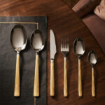 An elegant 36-piece stainless steel cutlery set with gold-finished floral engraved handles, arranged neatly on a dark placemat atop a wooden table, consisting of spoons, forks, and knives.