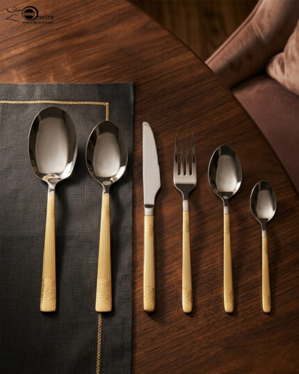 An elegant 36-piece stainless steel cutlery set with gold-finished floral engraved handles, arranged neatly on a dark placemat atop a wooden table, consisting of spoons, forks, and knives.