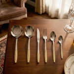 A complete 36-piece stainless steel cutlery set arranged on a dark wooden dining table, showcasing elegant gold-textured handles, including knives, forks, various spoons, and serving utensils.