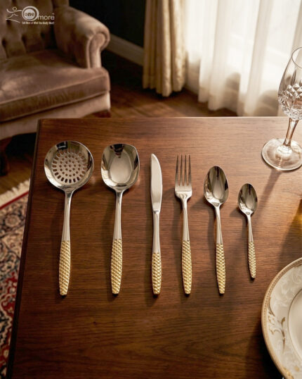 A complete 36-piece stainless steel cutlery set arranged on a dark wooden dining table, showcasing elegant gold-textured handles, including knives, forks, various spoons, and serving utensils.