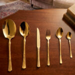 High-angle studio shot of a 36-piece gold-plated cutlery set arranged on a polished wooden surface, featuring matching serving pieces, dinner spoons, forks, knives, and teaspoons with elegant handle detailing.