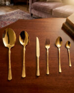 High-angle studio shot of a 36-piece gold-plated cutlery set arranged on a polished wooden surface, featuring matching serving pieces, dinner spoons, forks, knives, and teaspoons with elegant handle detailing.
