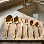 36-piece gold-finished cutlery set featuring delicate floral engraving on handles, arranged on a linen napkin, including table spoons, tea spoons, forks, knives, and serving utensils.