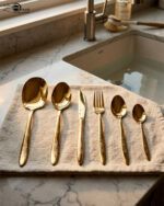 36-piece gold-finished cutlery set featuring delicate floral engraving on handles, arranged on a linen napkin, including table spoons, tea spoons, forks, knives, and serving utensils.