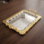 An elegant rectangular stainless steel serving tray with a highly reflective mirror finish and intricate center engravings, featuring a luxurious ornate gold border and decorative handles, displayed on a dark wooden table as shown in 1.jpg.