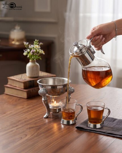 Luxury glass tea and coffee warmer carafe set with an ornate silver-plated burner stand, matching glass cup, and spoon on a polished wooden dining table.
