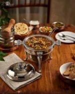 One More 4-liter glass chafing dish soup warmer with ornate stainless steel stand, ladle, and buffet burner on a wooden dining table.