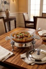Premium 2-liter glass chafing dish with an ornate silver stand, dome lid, and integrated buffet burner holder on a wooden dining table, branded by One More.