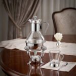 Luxury glass tea and coffee warmer carafe set with an ornate silver-plated burner stand, matching glass cup, and spoon on a polished wooden dining table.