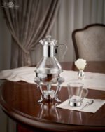 Luxury glass tea and coffee warmer carafe set with an ornate silver-plated burner stand, matching glass cup, and spoon on a polished wooden dining table.