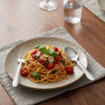 A fork and spoon from the 37-piece stainless steel cutlery set elegantly placed on a linen napkin next to a freshly served pasta dish.