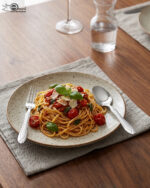 A fork and spoon from the 37-piece stainless steel cutlery set elegantly placed on a linen napkin next to a freshly served pasta dish.