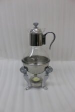 Luxury glass tea and coffee warmer carafe set with an ornate silver-plated burner stand, matching glass cup, and spoon on a polished wooden dining table.