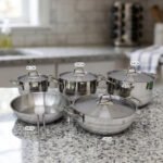 Five pieces of stainless steel cookware, including pots and a frying pan with glass lids, arranged on a granite countertop. Sizes 16cm, 20cm, and three 24cm pieces are clearly visible with price tags attached. Product features a non-stick interior and is exclusively available in Pakistan at OneMore.