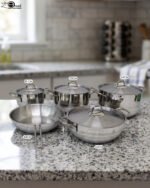 Five pieces of stainless steel cookware, including pots and a frying pan with glass lids, arranged on a granite countertop. Sizes 16cm, 20cm, and three 24cm pieces are clearly visible with price tags attached. Product features a non-stick interior and is exclusively available in Pakistan at OneMore.
