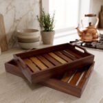 Two stacked, high-quality wooden serving trays resting on a kitchen countertop with a copper kettle and plant in the background.