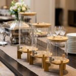Three glass beverage dispensers of varying heights, each featuring a bamboo lid and resting on a wooden stand, displayed on a table next to a buffet burner.