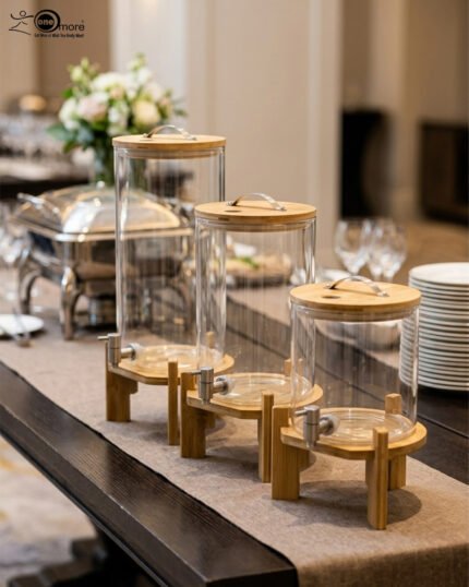 Three glass beverage dispensers of varying heights, each featuring a bamboo lid and resting on a wooden stand, displayed on a table next to a buffet burner.