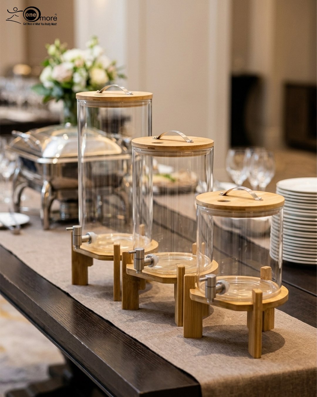1 Three glass beverage dispensers of varying heights, each featuring a bamboo lid and resting on a wooden stand, displayed on a table next to a buffet burner.