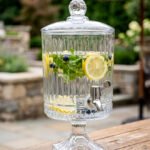 Elegant 4.5 Liter ribbed glass beverage dispenser with a silver spigot and decorative stand, filled with water, lemon slices, blueberries, and mint leaves on an outdoor wooden table, as shown in 2.jpg.