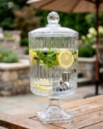 Elegant 4.5 Liter ribbed glass beverage dispenser with a silver spigot and decorative stand, filled with water, lemon slices, blueberries, and mint leaves on an outdoor wooden table, as shown in 2.jpg.