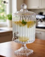 Elegant 4.5L glass beverage dispenser filled with iced lemonade and mint, resting on a pedestal stand on a wooden kitchen counter.