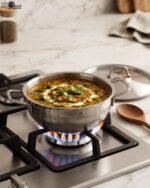 Five pieces of stainless steel cookware, including pots and a frying pan with glass lids, arranged on a granite countertop. Sizes 16cm, 20cm, and three 24cm pieces are clearly visible with price tags attached. Product features a non-stick interior and is exclusively available in Pakistan at OneMore.