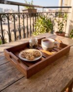 Two stacked, high-quality wooden serving trays resting on a kitchen countertop with a copper kettle and plant in the background.