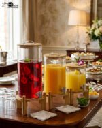 Three glass beverage dispensers of varying heights, each featuring a bamboo lid and resting on a wooden stand, displayed on a table next to a buffet burner.