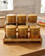 A set of six glass spice jars with wooden-look lids arranged on a matching tiered wooden stand, sitting on a kitchen countertop.