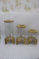 Three glass beverage dispensers of varying heights, each featuring a bamboo lid and resting on a wooden stand, displayed on a table next to a buffet burner.