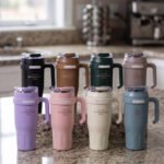 : An array of colorful 1.2L stainless steel tumblers with handles and clear lids displayed on a kitchen counter