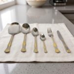 Six-piece gold accented stainless steel flatware set displayed on a linen mat including rice spoons, fork, knife, and serving spoons.