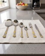 Six-piece gold accented stainless steel flatware set displayed on a linen mat including rice spoons, fork, knife, and serving spoons.