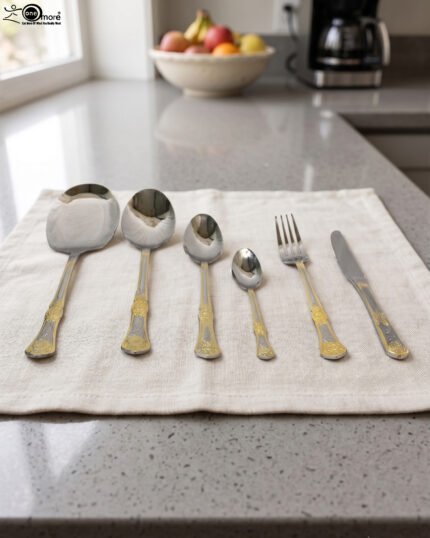 Six-piece gold accented stainless steel flatware set displayed on a linen mat including rice spoons, fork, knife, and serving spoons.