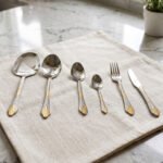 Six-piece luxury stainless steel cutlery set featuring textured handles with gold filigree details displayed on a beige linen napkin.