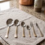 Gold-embossed stainless steel cutlery set including spoons, fork, and knife arranged on a beige cloth table setting.