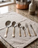 Gold-embossed stainless steel cutlery set including spoons, fork, and knife arranged on a beige cloth table setting.