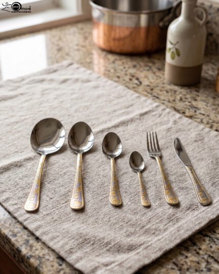 Gold-embossed stainless steel cutlery set including spoons, fork, and knife arranged on a beige cloth table setting.