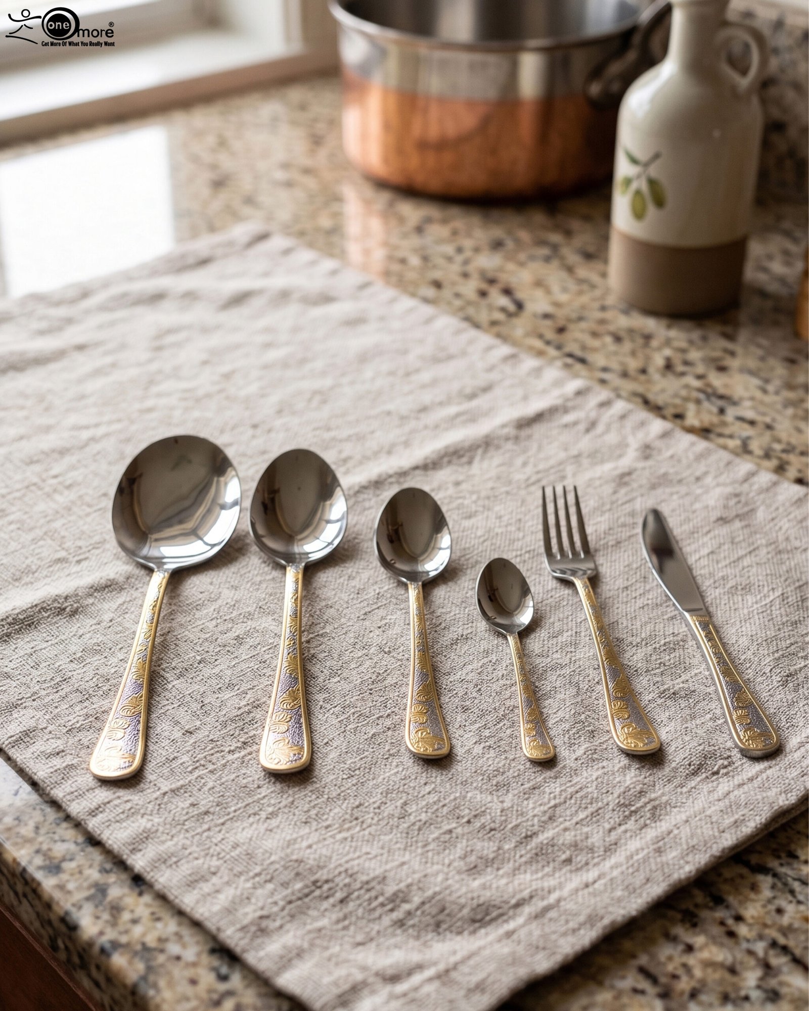 1 Gold-embossed stainless steel cutlery set including spoons, fork, and knife arranged on a beige cloth table setting.