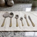 Stainless steel cutlery set with gold patterned handles featuring serving spoons, dinner forks, knives, and dessert spoons on a linen cloth.
