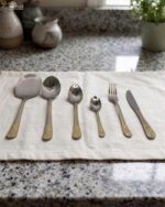 Stainless steel cutlery set with gold patterned handles featuring serving spoons, dinner forks, knives, and dessert spoons on a linen cloth.