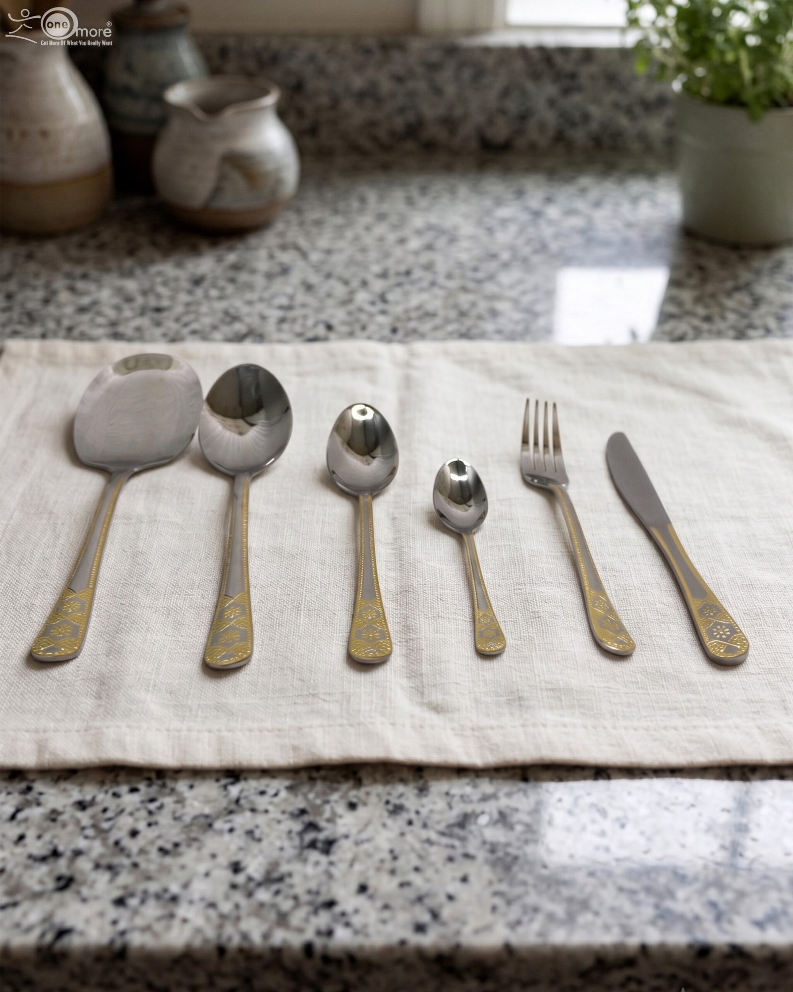 1 Stainless steel cutlery set with gold patterned handles featuring serving spoons, dinner forks, knives, and dessert spoons on a linen cloth.