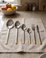 Stainless steel cutlery set with textured gold handles and floral engravings displayed on a beige cloth.