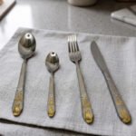 24-piece stainless steel cutlery set featuring gold "Love" engravings on handles, including rice spoon, sweet spoon, fork, and knife on a gray napkin.