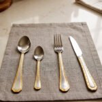 24-piece stainless steel cutlery set with gold-bordered handles including a rice spoon, sweet spoon, fork, and knife arranged on a grey placemat.