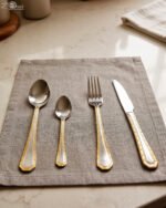 24-piece stainless steel cutlery set with gold-bordered handles including a rice spoon, sweet spoon, fork, and knife arranged on a grey placemat.