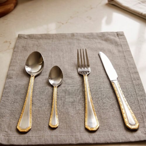 24-piece stainless steel cutlery set with gold-bordered handles including a rice spoon, sweet spoon, fork, and knife arranged on a grey placemat.