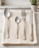 24-piece stainless steel cutlery set with ornate gold embossed handles laid out on a beige linen napkin